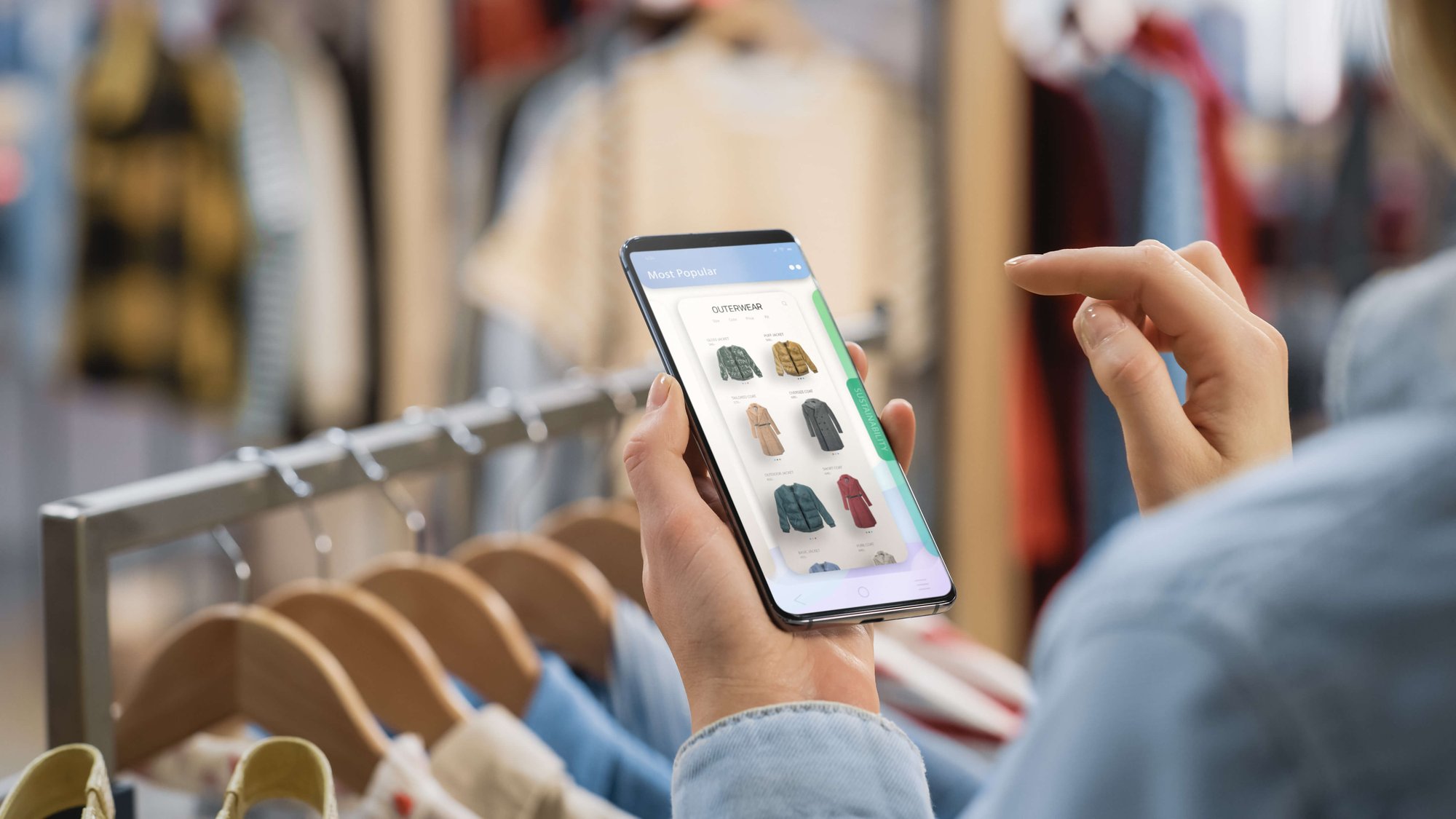 Frau hält Smartphone mit Benutzeroberfläche eines Online-Modehauses in einem Kleidungsgeschäft