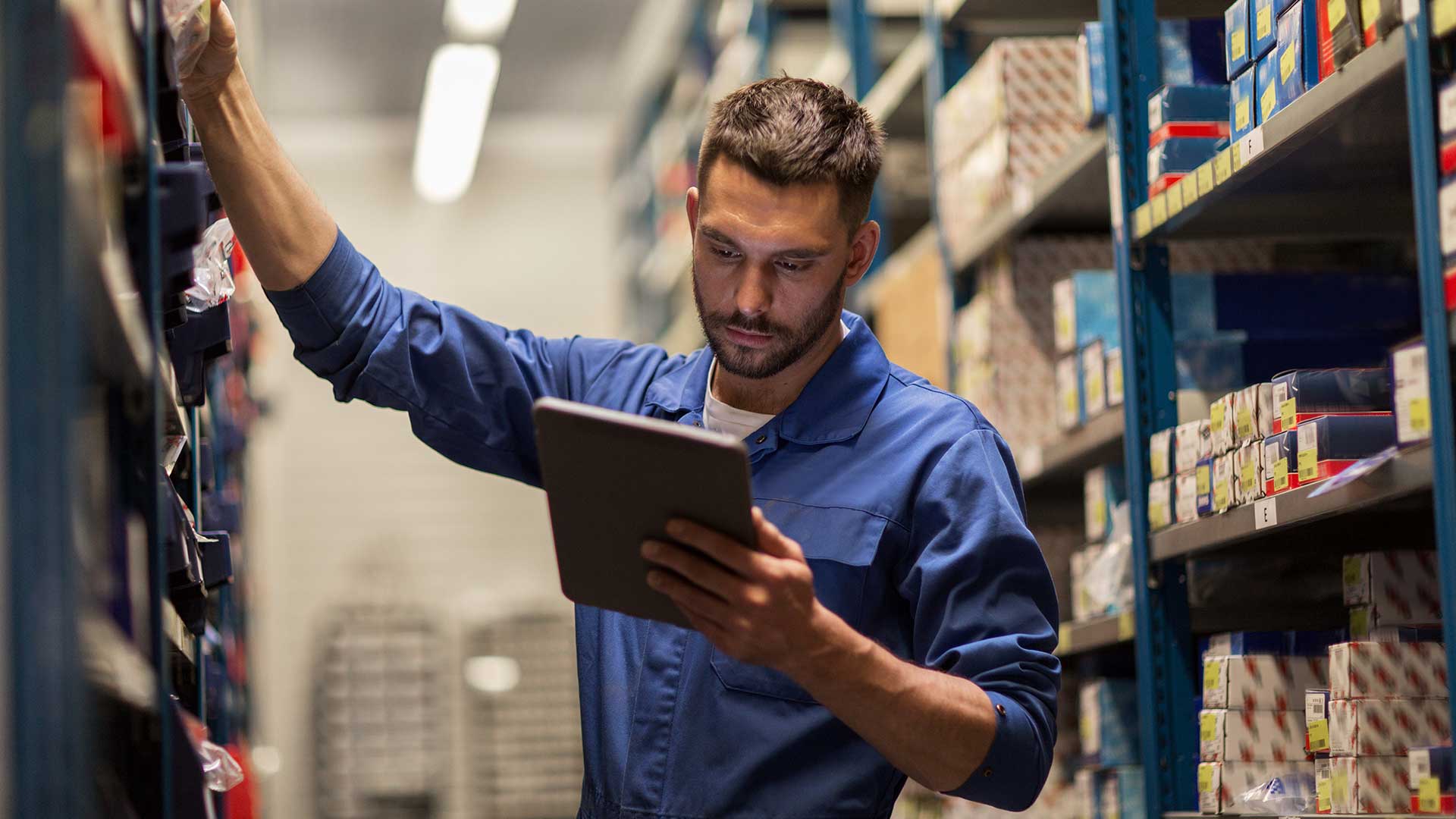 A man in overalls stands at a shelf in the warehouse with a tablet in his hand